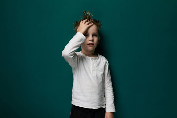 Ένα νεαρό αγόρι στέκεται με τα χέρια του στο κεφάλι του σε ένα ζωντανό πράσινο τοίχο. Εμφανίζεται βαθιά στη σκέψη ή την περισυλλογή, με ένα απλό και μινιμαλιστικό υπόβαθρο. - Φωτογραφία, εικόνα