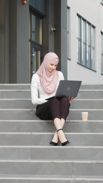 Verticaal scherm: Jonge moslim vrouw zit op de trap in de buurt van business center en werkt aan draadloze laptop. Mooie vrouw met elegante kleren en hijab. Freelance concept. Moslimvrouw aan het werk. - Video