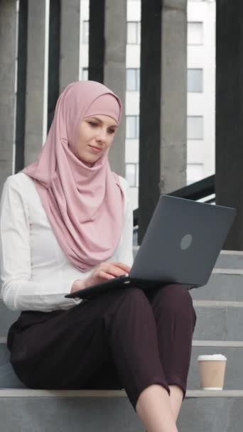 Vertikální obrazovka: Okouzlující žena ve stylovém elegantním oblečení a hidžábu. Muslimka pracující na dálku. Portrét atraktivní arabské ženy v elegantním oblečení a hidžábu sedící na schodech s moderním - Záběry, video