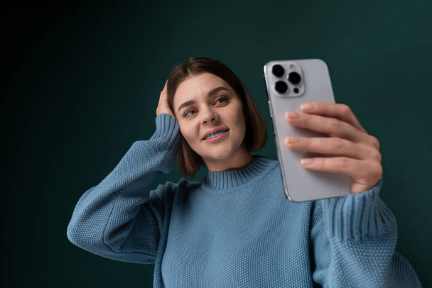 Egy nő, aki egy telefont tart az arca előtt, mosolyog és egy szelfihez pózol. Az elülső kamerát használja, hogy rögzítse magát abban a pillanatban.. - Fotó, kép