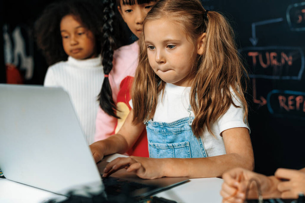 Inteligentna dziewczyna w białym śliniaczku uczy się kodowania technologii robotyki przy użyciu laptopa w klasie STEM. Uczeń w niebieskiej koszuli stara się edukować płytę główną, podczas gdy inteligentne uczennice czytają kod. Erudycja. - Zdjęcie, obraz