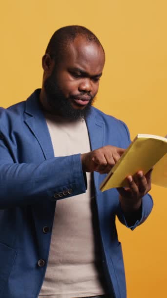 Vertical video Man struggling to read book with small font, pointing finger to words on page. Person enjoying literature novel, wetting fingers to change page, isolated over studio background, camera - Footage, Video