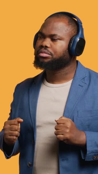 Függőleges videó Boldog afro-amerikai személy szórakozik, elektronikus dalokat hallgat fejhallgatón keresztül. Vidám BIPOC úgy tesz, mintha DJ, élvezi a tánc zene, stúdió háttér, kamera A - Felvétel, videó