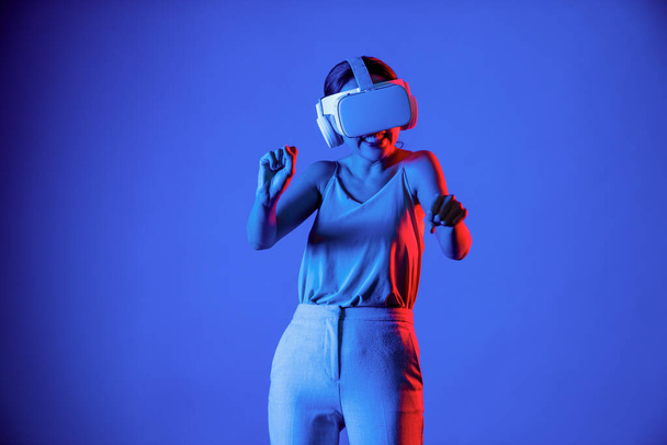 Smart female stand surrounded by neon light wearing VR headset connecting metaverse, future cyberspace community technology. Elegant woman enjoy playing car racing games in meta world. Hallucination. - Photo, Image