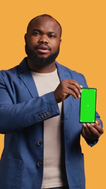 Κάθετο βίντεο Χαρούμενος Αφροαμερικάνος που συστήνει πράσινο smartphone με οθόνη, γέλιο, φόντο στούντιο. Joyous BIPOC πρόσωπο παρουσιάζει mockup κινητό τηλέφωνο, δίνοντας θετική ανατροφοδότηση, κάμερα Β - Πλάνα, βίντεο
