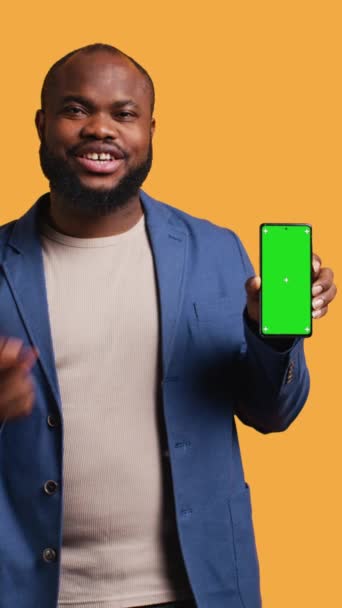 Video verticale Uomo afroamericano sorridente che presenta il cellulare con display a schermo verde, isolato sullo sfondo dello studio. Allegro persona BIPOC creazione di promozione con spazio vuoto copia mockup telefono - Filmati, video