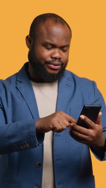 Vertical video Portrait of energetic happy man holding smartphone celebrating, throwing fists in air from excitement. Overjoyed BIPOC person reading good news on phone, cheering, studio background - Footage, Video