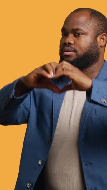 Pionowe wideo Portret wesołego, przyjaznego Afrykańczyka wykonującego gestem symbolicznego serca. Wesoła, opiekuńcza osoba BIPOC pokazująca gest miłości, odizolowana od tła studia - Materiał filmowy, wideo