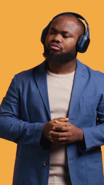 Vertical video Relaxed african american man having fun listening to music on headphones during time off. Carefree BIPOC person enjoying songs and singing lyrics, isolated over studio background - Footage, Video