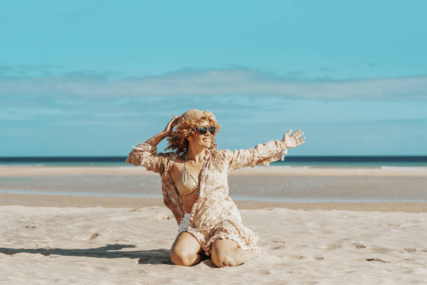 Vista frontal de una hermosa mujer con los brazos abiertos sentada en la playa. Mujer rubia con el pelo rizado soplando en el viento se relaja mirando y sonriendo. - Foto, imagen