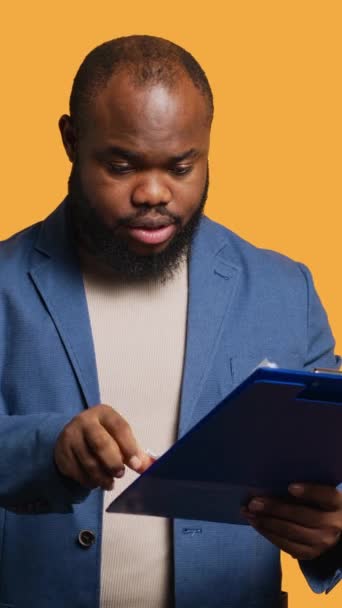 Pionowe wideo Człowiek przeglądając sprawozdanie finansowe z audytu na schowku, zadowolony przez wysokie dochody, tło studio. Szczęśliwy afrykański pracownik czytający papiery na notatniku, zadowolony z danych liczbowych - Materiał filmowy, wideo