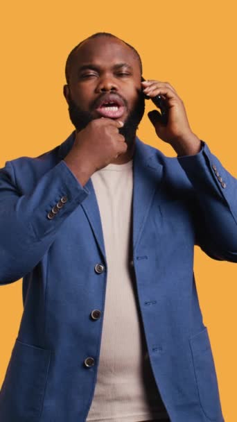 Κατακόρυφο βίντεο Αφρο-αμερικάνος που έχει στοχαστική συνομιλία με φίλο κατά τη διάρκεια τηλεφωνικής κλήσης, στούντιο φόντο. Ψυχαναγκαστικό άτομο της BIPOC να μιλάει στο τηλέφωνο με τον σύντροφο, να σκέφτεται να κάνει χειρονομίες - Πλάνα, βίντεο