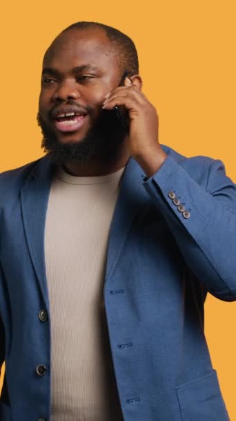 Függőleges videó Örömteli ember pozitív érzelmekkel, élvezi a beszélgetést barátaival telefonhívás segítségével okostelefon, stúdió háttér. Boldog afro-amerikai személy mosollyal az arcán telefonhívás közben - Felvétel, videó