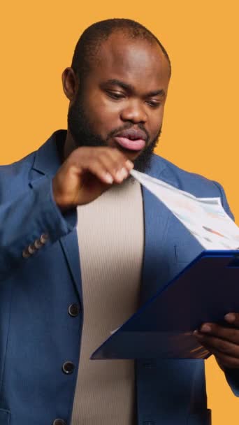 Вертикальне відео Чоловік переглядає, хоча фінансовий аудит звіт на кишені, позіхання, відчуття втоми, студійний фон. Набридлий афроамериканський працівник виснажений під час читання паперу на блокноті, камера А - Кадри, відео