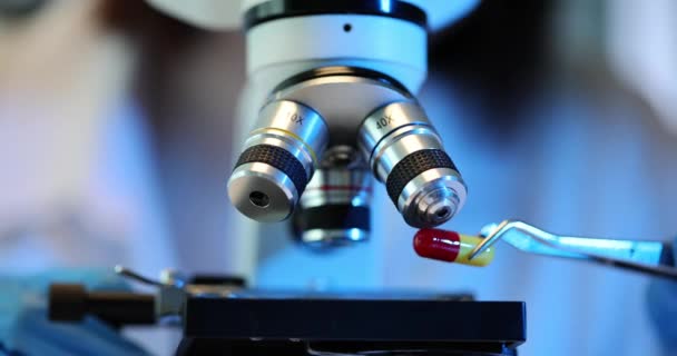 Onderzoeker plaatst capsule met pincet voor kwaliteitsanalyse onder microscoop. Laboratoriummedewerker houdt zich bezig met het bestuderen van pil met behulp van moderne apparatuur - Video