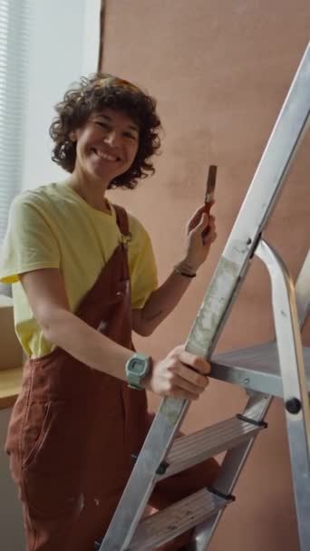 Imágenes de POV verticales de traer rodillo de pintura a la mujer caucásica que pinta paredes usando escalera y engaño durante la renovación del hogar - Imágenes, Vídeo