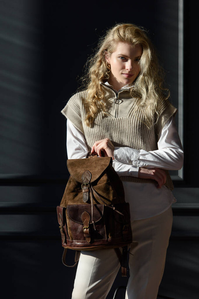 A woman holds brown leather backpack. Model wearing stylish knitted vest, white shirt and classic trousers. - Photo, Image