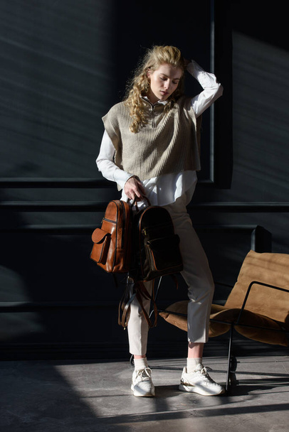 Una mujer sostiene la mochila de cuero marrón. Modelo con elegante chaleco de punto, camisa blanca y pantalones clásicos. - Foto, imagen