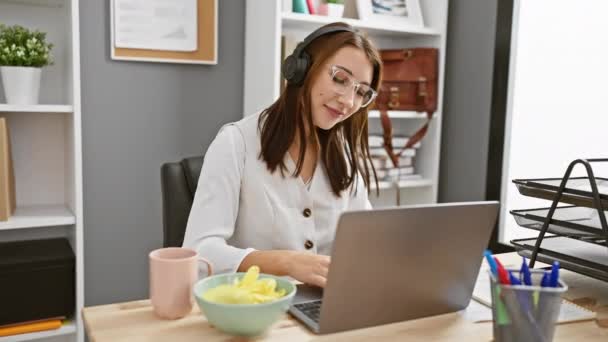 Mladá brunetka žena pracuje pozorně na notebooku v moderní kanceláři, zatímco nosí sluchátka a jíst občerstvení - Záběry, video