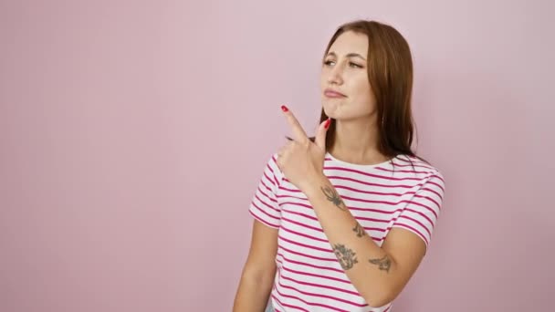 Giovane ragazza bruna indossa strisce maglietta allegra con un sorriso sul viso che punta con mano e dito verso il lato con espressione felice e naturale su sfondo rosa isolato - Filmati, video
