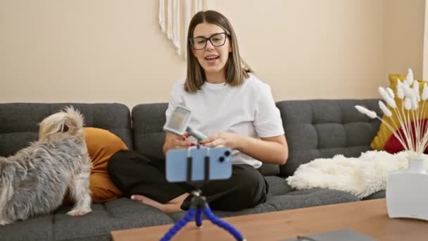 Ισπανίδα γυναίκα με γυαλιά vlogging στο σπίτι με το σκύλο της, χρησιμοποιώντας smartphone σε τρίποδο, στο άνετο καθιστικό ρύθμιση. - Πλάνα, βίντεο