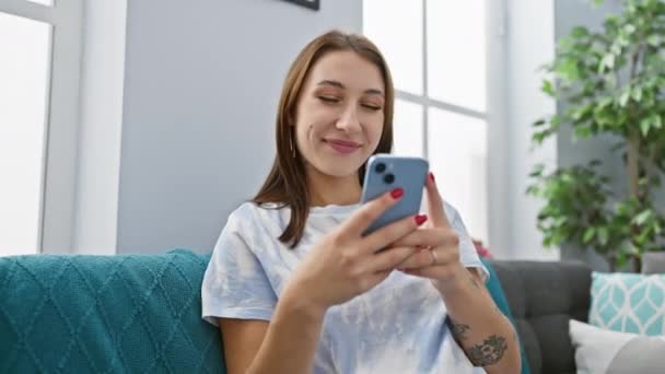 Μια χαρούμενη νεαρή γυναίκα απολαμβάνει να χρησιμοποιεί το smartphone της σε έναν άνετο καναπέ σε εσωτερικούς χώρους, ενσωματώνοντας έναν χαλαρό τρόπο ζωής στο σπίτι. - Πλάνα, βίντεο