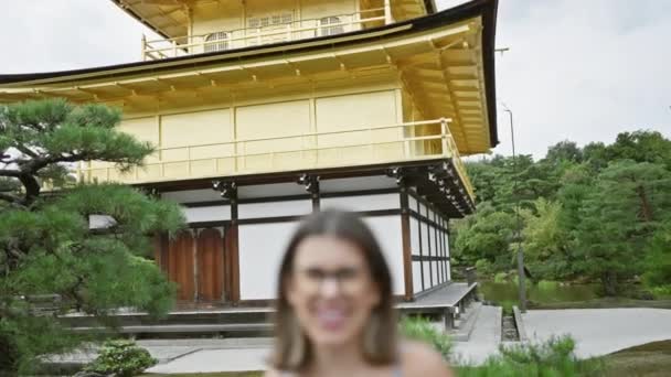 Приваблива іспаномовна жінка, радість, що випромінюється від її мерехтливої посмішки, стоїть впевнено і випадково в її окулярах на захоплюючому золотому павільйоні Кінкаку-дзі, коштовності Кіото, Японія - Кадри, відео