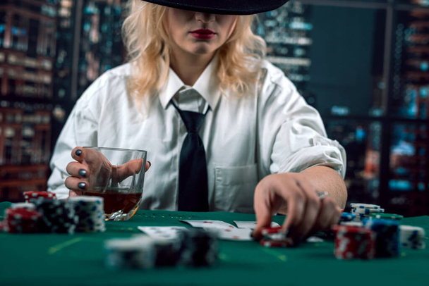 ελκυστικό κορίτσι με πουκάμισο και καπέλο πίνοντας ουίσκι και κοιτάζοντας κάρτες πόκερ στο καζίνο. Ο γυναικάς βάζει στοίχημα. πόκερ στο καζίνο - Φωτογραφία, εικόνα