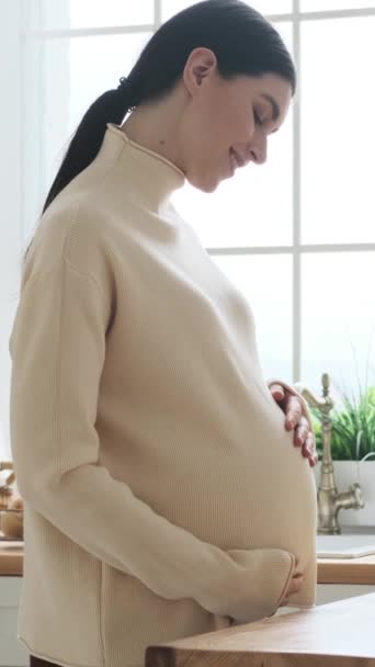 Vrolijke en vrolijke blanke zwangere vrouw raakt buik thuis keuken. Zwangerschap, baby en bevalling verwachten concept. Verticale video. - Video