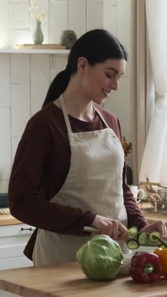 Ευτυχισμένη και θετική λευκή γυναίκα που φοράει ποδιά και ετοιμάζει φρέσκια σαλάτα λαχανικών στο σπίτι. Υγιεινά τρόφιμα και μαγειρική έννοια. Κάθετη βίντεο. - Πλάνα, βίντεο