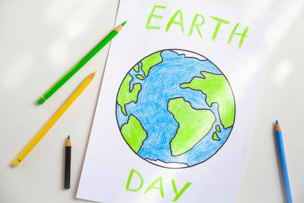 Una hoja blanca de papel que representa un globo pintado con lápices de colores y la inscripción Día de la Tierra está en la mesa iluminada por la luz del día. - Foto, Imagen