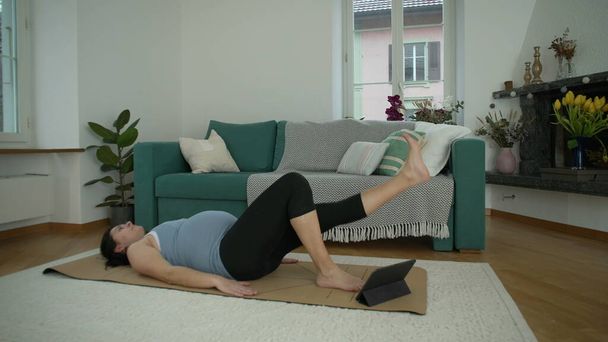 Üçüncü üç aylık hamilelik döneminde evde egzersiz yapan kadın oturma odasında uzanıp bacağını kaldırıyordu. Hamilelik öncesi egzersizleri sırasında 30 'lu yaşlarda kadın sporu. - Fotoğraf, Görsel