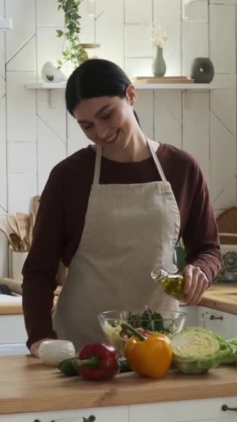 Щаслива кавказька жінка засолює і перемішує свіжий салат у мисці, використовуючи дерев'яну ложку на домашній кухні. Приготування здорової їжі. Вертикальне відео. - Кадри, відео