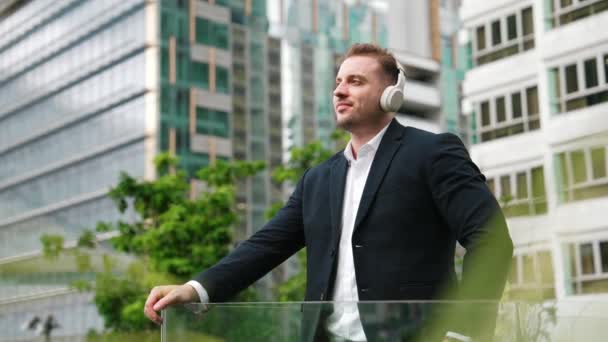 Manager mit Kopfhörer hören gerne Musik, während sie auf dem Dach stehen. Entspannt trägt der Investor ein Headset und bewegt sich zum Musikreim, während er auf den modernen Wolkenkratzer blickt. Lebensstil. Urbane Pflanzen. - Filmmaterial, Video