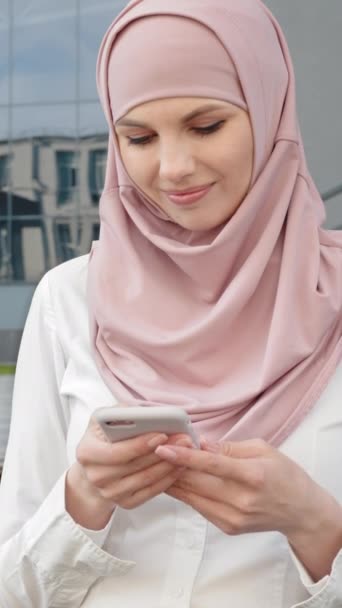Pantalla vertical: mujer musulmana usando teléfono inteligente mientras trabaja en el ordenador portátil. Mujer agradable en hijab sentado en el banco de madera al aire libre y la celebración de teléfono celular moderno. - Imágenes, Vídeo