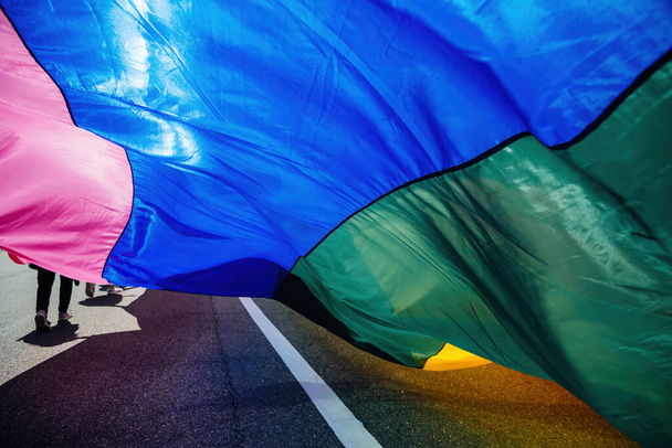Vibrante grande bandiera dell'orgoglio ondeggia nella brezza durante una parata, con le gambe dei popoli visibili sotto - Foto, immagini