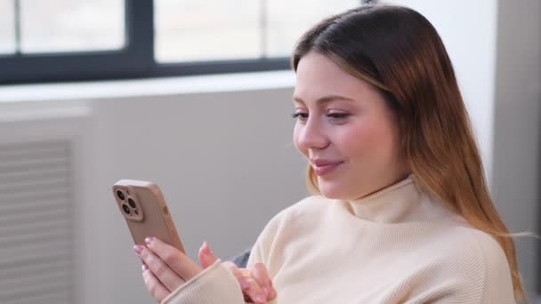 Портрет щасливої кавказької молодої жінки, яка розслабляється онлайн з телефоном вдома. Повідомлення в додатку соціальних мереж, читання новин, перегляд Інтернету. - Кадри, відео