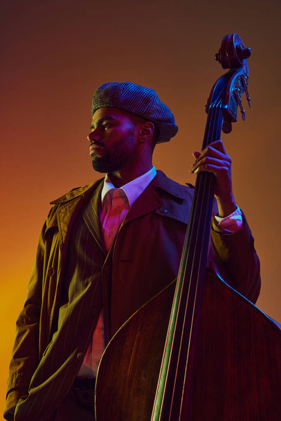 Retrato de homem afro-americano, jogador com expressão serena posando com instrumento de corda em luz de néon contra fundo gradiente. Conceito de arte, música, música clássica e estilo de vida moderno. Anúncio - Foto, Imagem