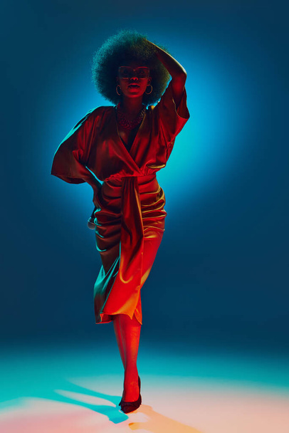 Элегантная афроамериканка со стильной прической в ретро-платье, танцующая в неоновом свете на градиентном фоне. Концепция искусства, музыки, хобби, классической музыки и современного образа жизни - Фото, изображение