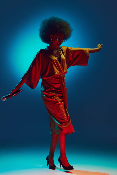 Mujer afroamericana en vestido de estilo retro bailando mientras canta en el escenario en focos de neón contra el fondo degradado. Concepto de arte, música, hobby, música clásica y estilo de vida moderno. Anuncio - Foto, Imagen