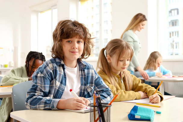 Glücklicher Junge, der am Schreibtisch neben seinem Klassenkameraden sitzt, während der Lehrer den Schülern im Hintergrund hilft. Bildung, Lernen und Menschen-Konzept - Foto, Bild