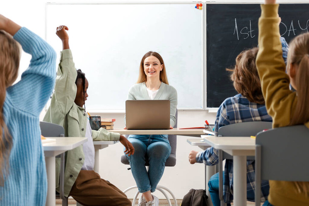 ハッピー先生が小学生に小学校でノートパソコンを使って机の上に座って質問をする. 教育,学習,人々のコンセプト - 写真・画像