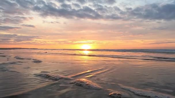 Esta grabación en cámara lenta captura la tranquila belleza de una puesta de sol en una playa serena, con suaves olas golpeando la orilla. El sol dorado se sumerge hacia el horizonte, reflejando sus tonos cálidos en - Metraje, vídeo