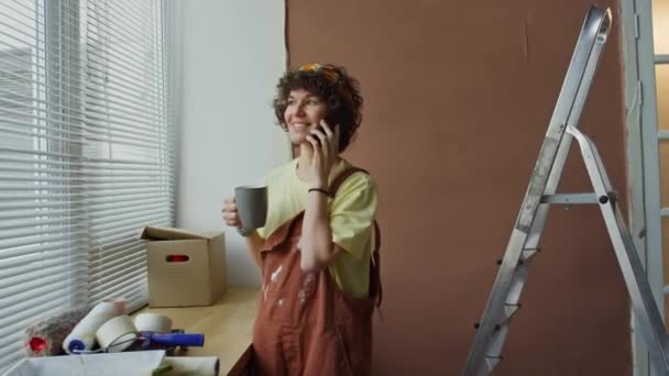 Imágenes laterales de la alegre mujer caucásica en mono teniendo conversación telefónica durante el café o el té de pie junto a la ventana en el hogar renovado - Imágenes, Vídeo