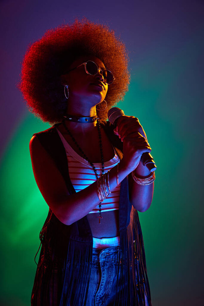 Young vocalist with iconic 70s afro, singing, dressed in black vest and beaded necklaces in neon light against gradient background. Concept of art, music, hobby, classical music and modern lifestyle. - Foto, afbeelding