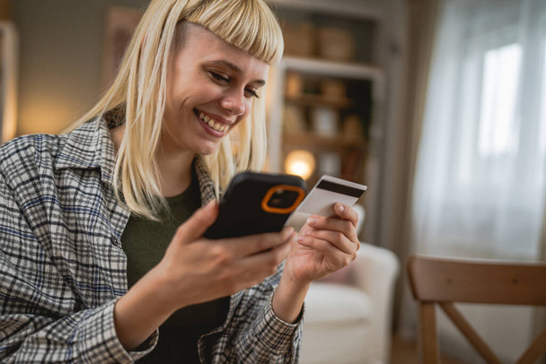 Jovem caucasiana usando telefone inteligente e compras on-line, segurando cartão de crédito fazendo o pagamento sentado na mesa em casa - Foto, Imagem