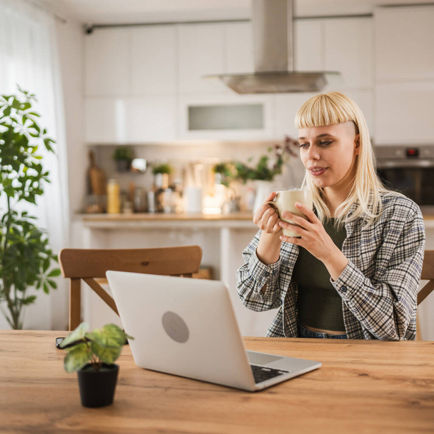 młody biały blondynka kobieta praca na laptop i trzymać kubek z w domu szczęśliwy pozytywny - Zdjęcie, obraz