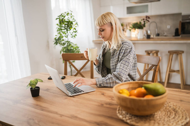 Молода біла жінка працює на ноутбуці і тримає чашку з дому щасливим позитивним - Фото, зображення