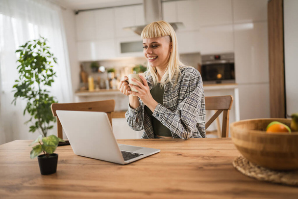 Jeune femme blonde caucasienne travailler sur ordinateur portable et tenir la tasse de la maison heureux positif - Photo, image