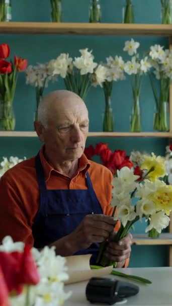 70 yaşında yaşlı bir çiçekçi çiçekçi çiçekçi dükkanında bir buket güzel bahar çiçeği düzenliyor. Dikey video - Video, Çekim